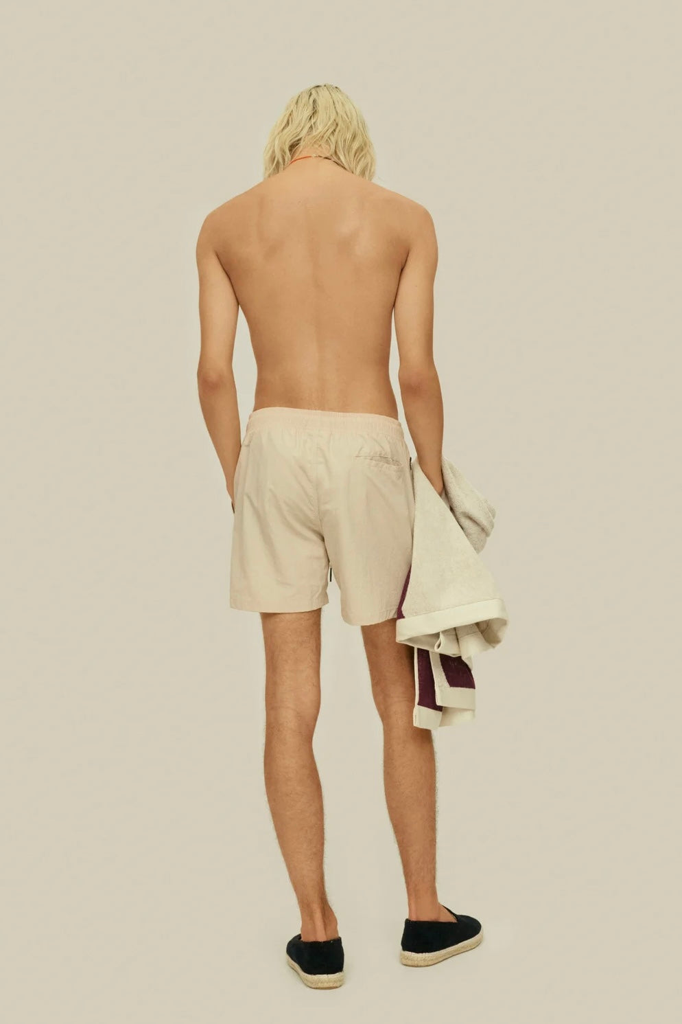 A shirtless man with long blonde hair, wearing beige shorts, seen from the back, holding a towel and magazines.