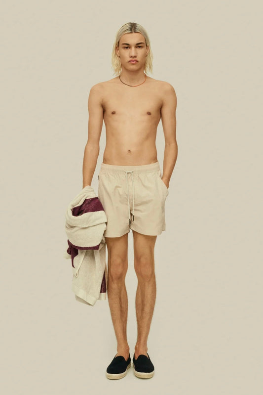 A young man standing shirtless in swim shorts, holding a towel, against a neutral background.
