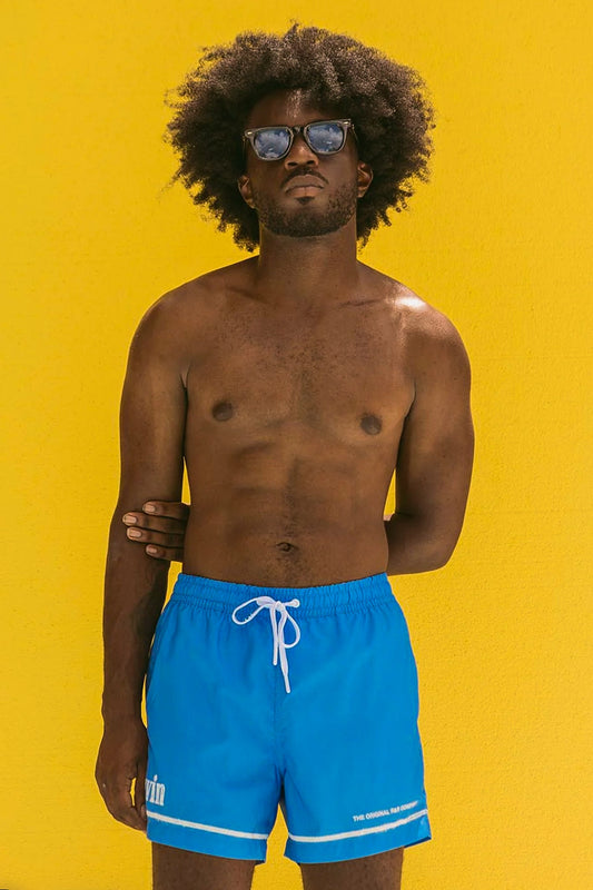 Man wearing blue swim trunks and sunglasses standing against a yellow background