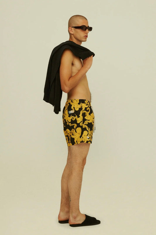 Side profile of a shirtless man wearing patterned shorts and sunglasses, holding a jacket over his shoulder.